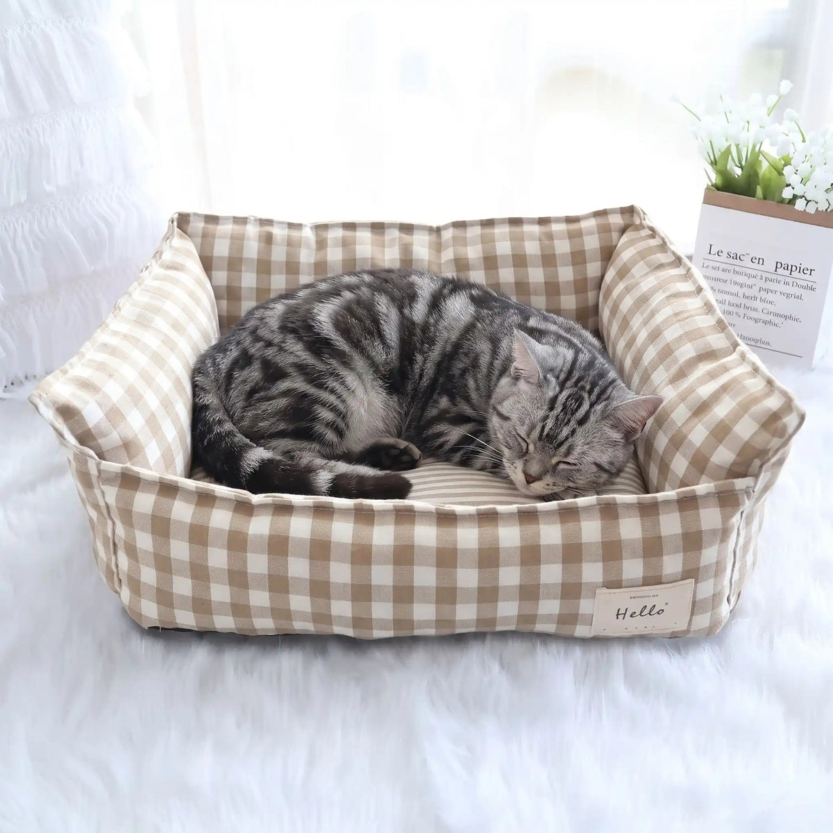 panier pour chat avec motif de carreaux de couleur abricot et blanc avec une beau chat gris et noir qui dort paisiblement dedans et le panier est en forme de sofa